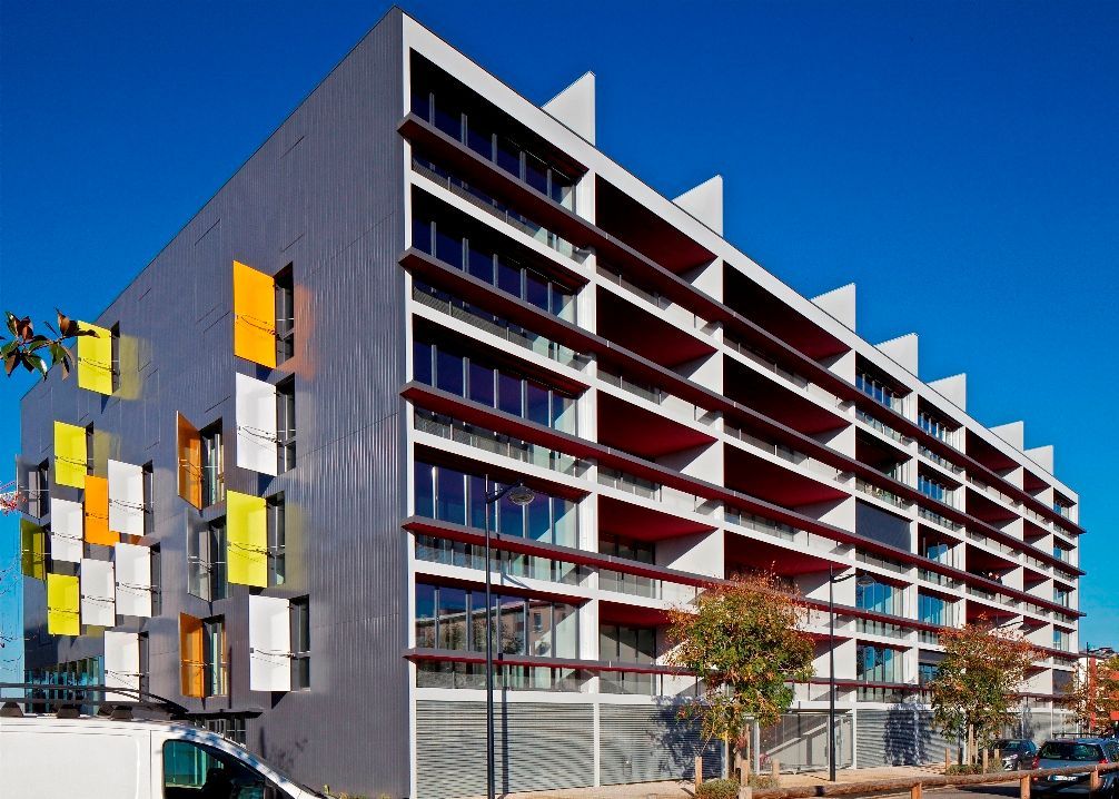 Bâtiment moderne avec façade en tôle ondulée, parasols rouges et volets colorés. Journée ensoleillée, ciel bleu.