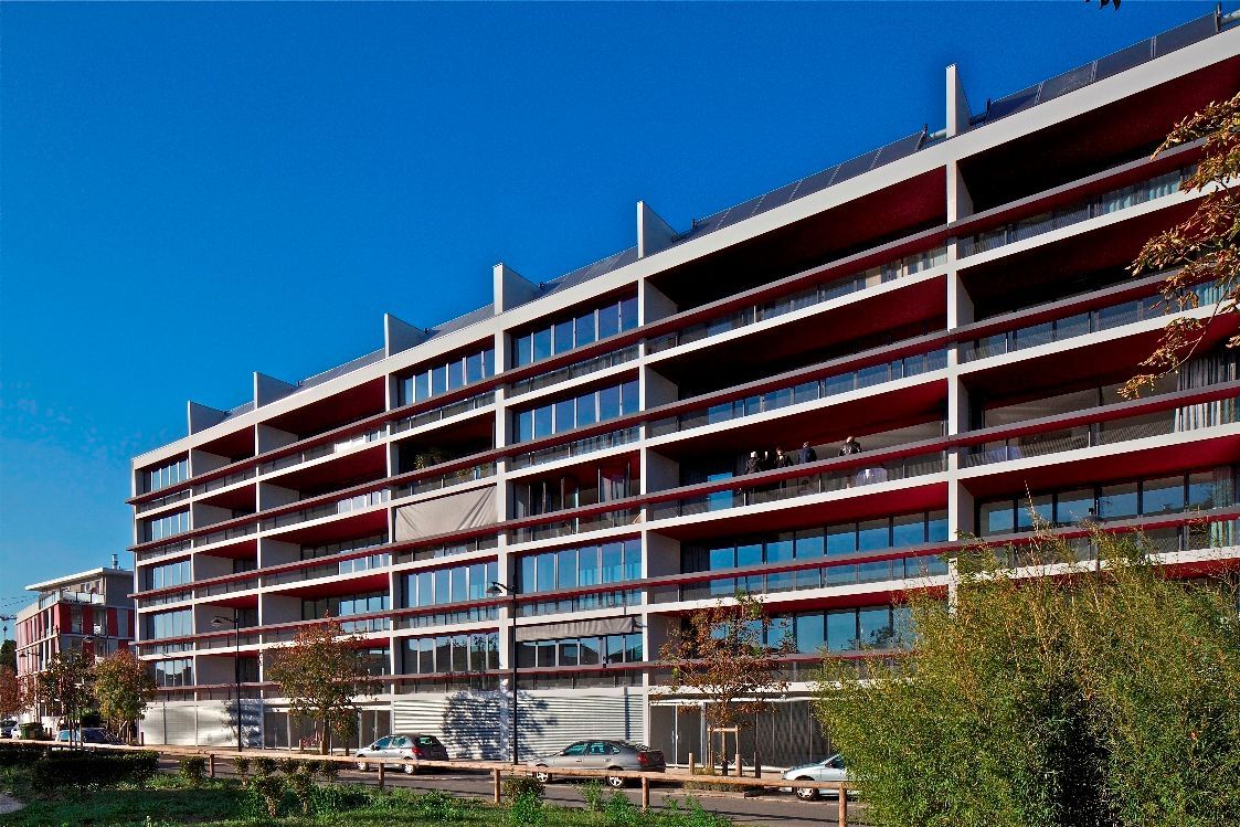 Immeuble d'appartements à plusieurs étages avec balcons rouges, sous un ciel bleu, vu depuis une zone herbeuse.