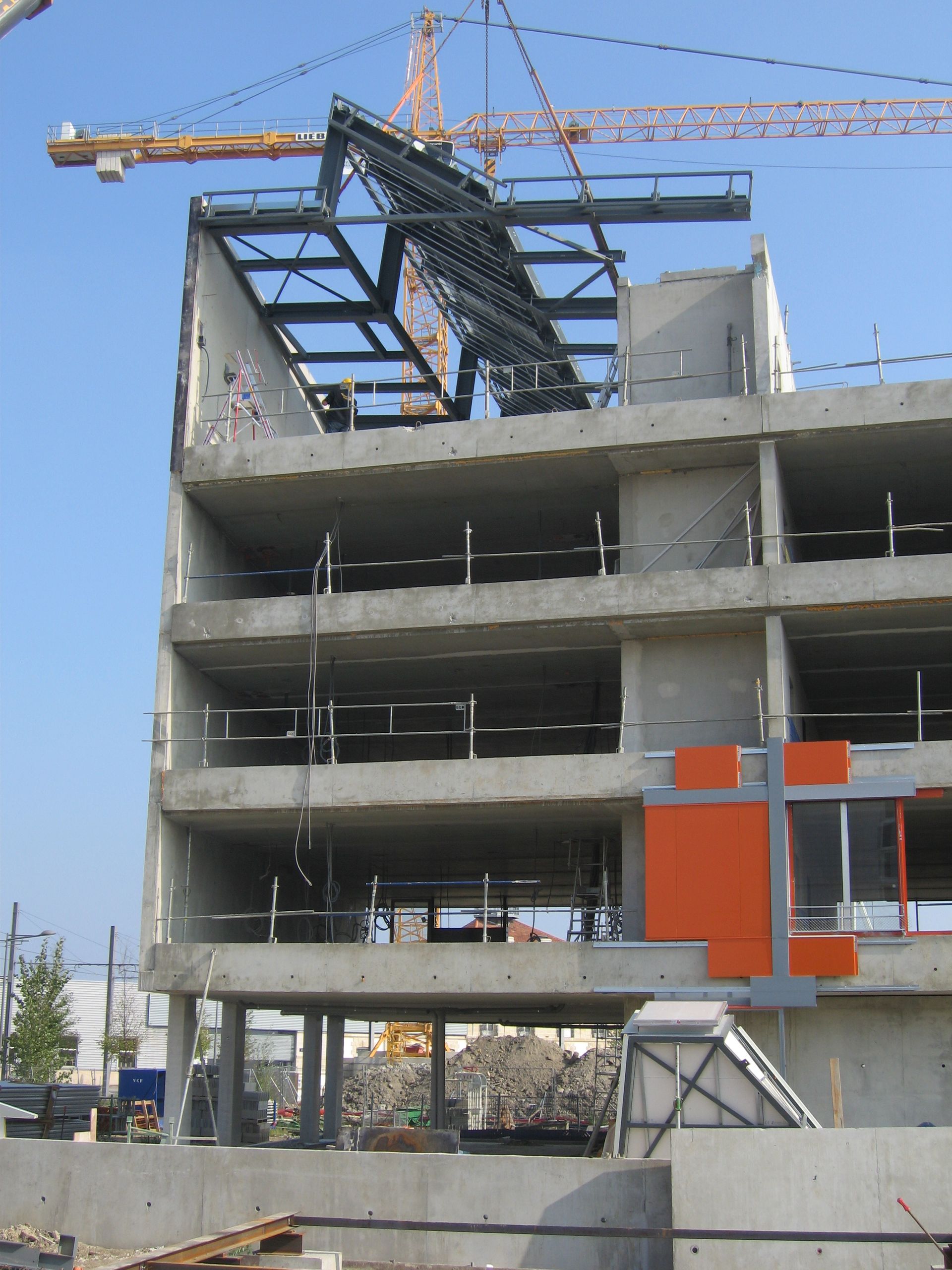 Chantier : bâtiment en béton en construction, structure en acier soulevée par grue.