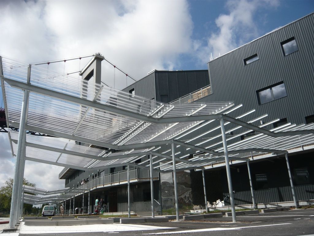 Auvent métallique au-dessus du parking devant un bâtiment moderne par une journée partiellement nuageuse.