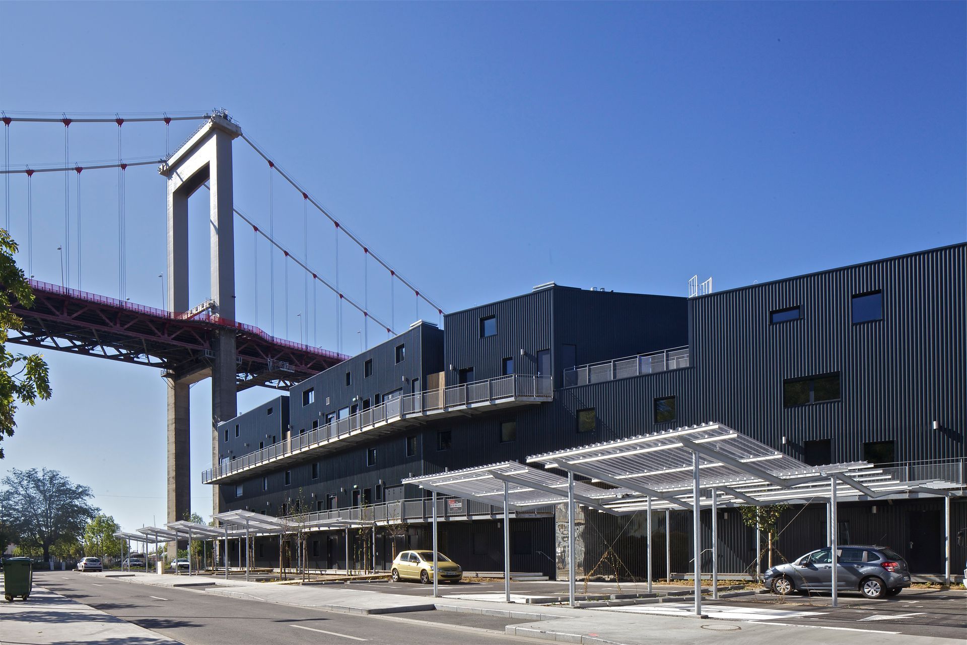 Bâtiment moderne avec panneaux solaires, sous un pont suspendu, ciel bleu.