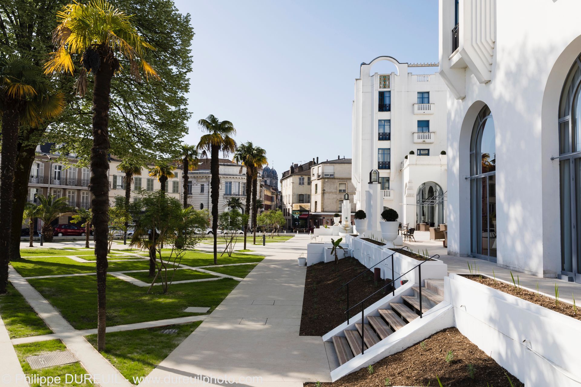 Vue d'une place extérieure ensoleillée avec des bâtiments blancs et des palmiers ; un escalier mène vers le bas.