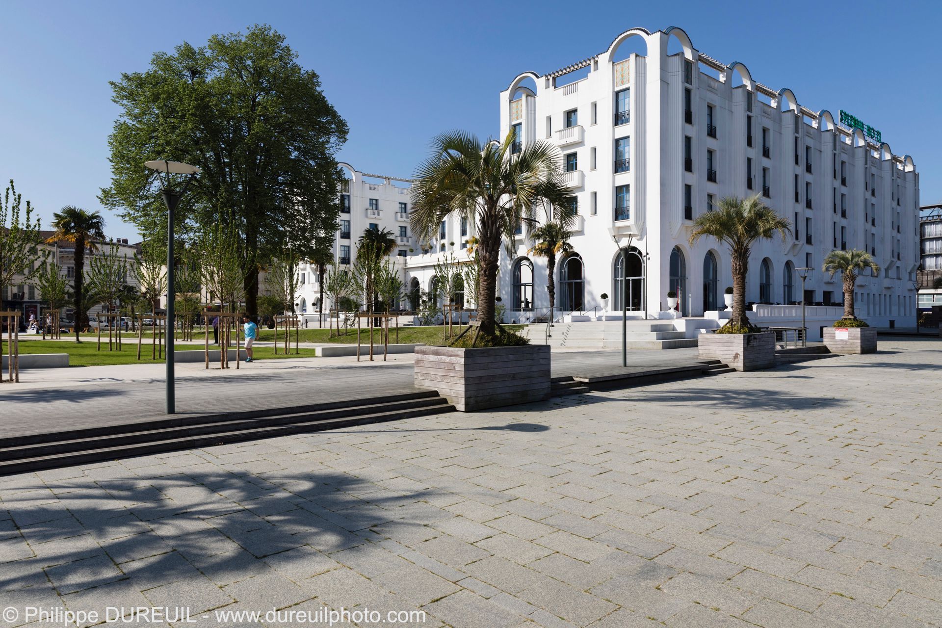 Hôtel blanc avec palmiers et place par une journée ensoleillée.