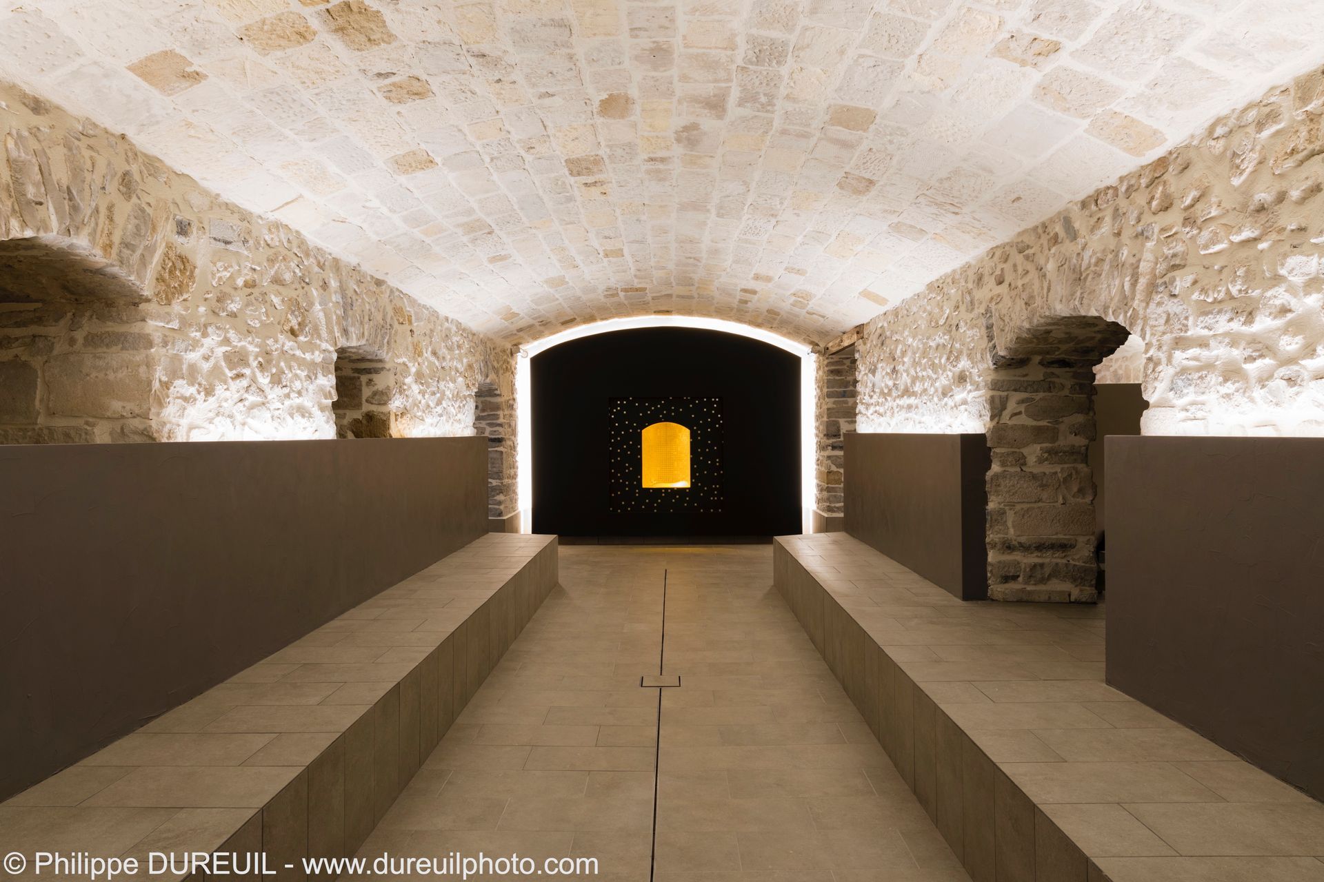 Cave en pierre avec un long couloir, des panneaux bruns et une porte noire avec une lumière jaune centrale.