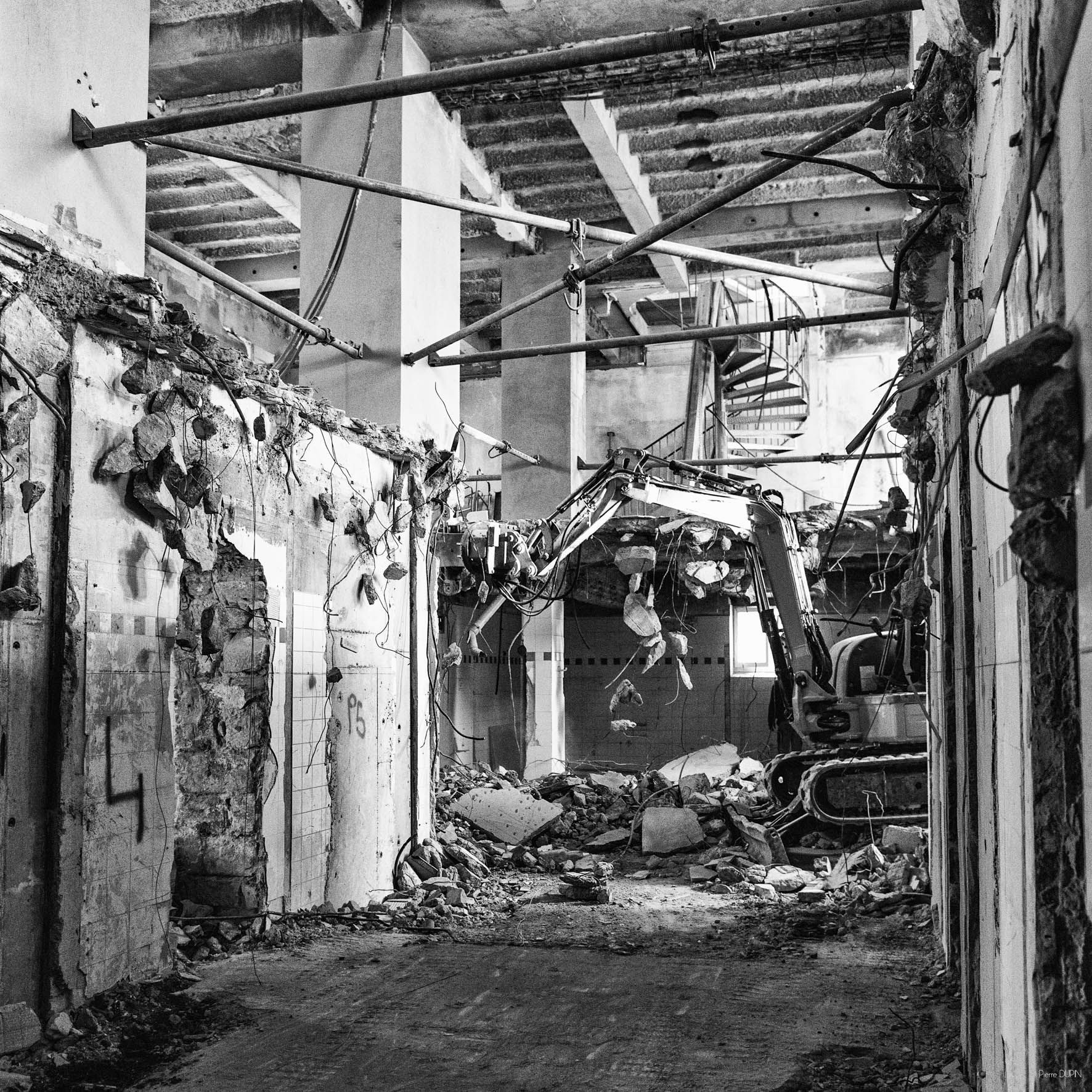 Démolition de l'intérieur d'un bâtiment, montrant une excavatrice travaillant dans un couloir avec des poutres et des débris apparents.