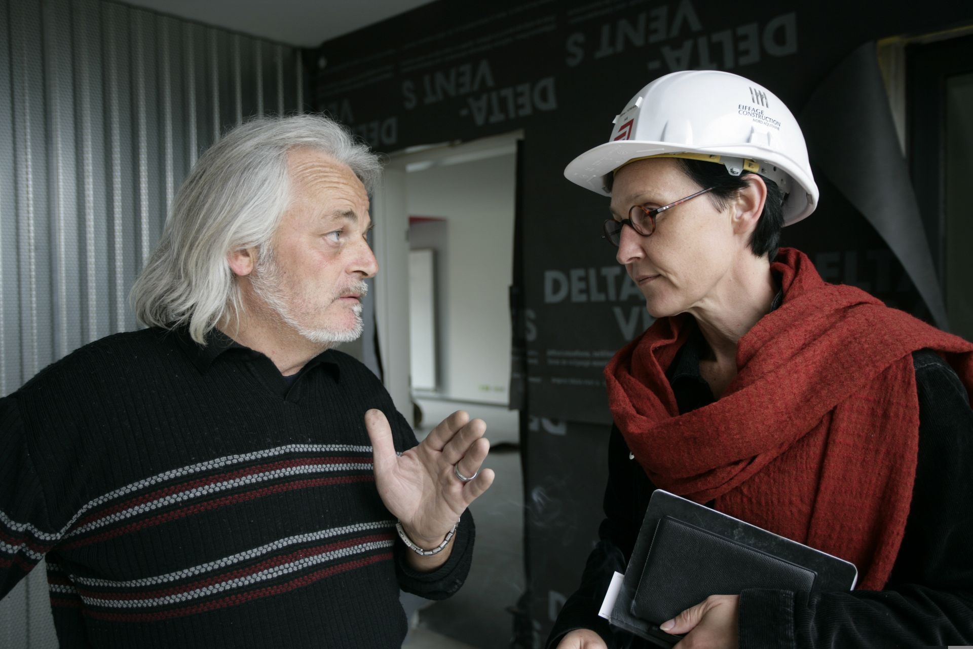 Un homme fait un geste et parle à une femme portant un casque de chantier, tous deux à l'intérieur d'un bâtiment en construction.