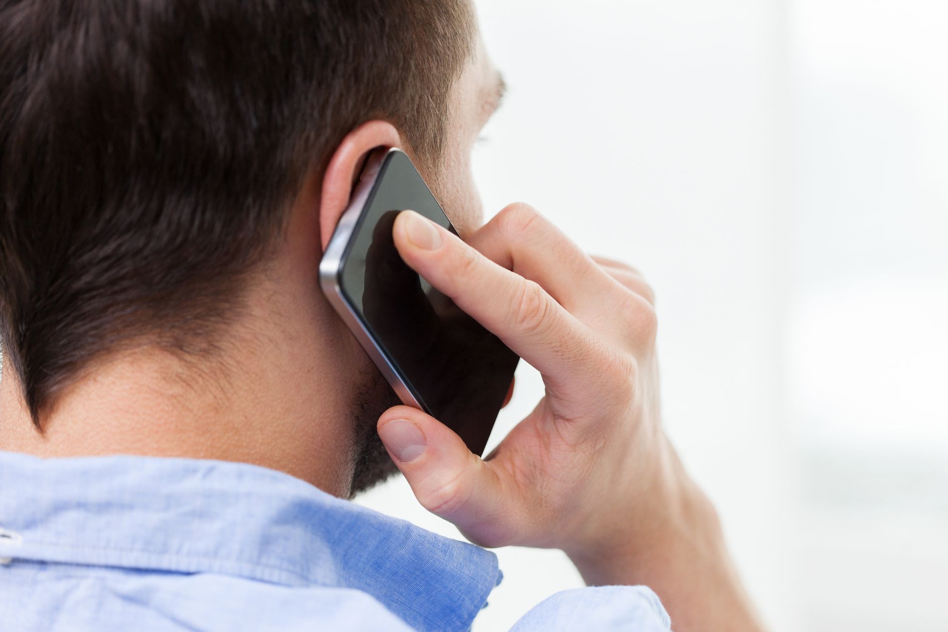 Eine Person, die ein Smartphone ans Ohr hält, ist von hinten zu sehen und trägt ein hellblaues Hemd.