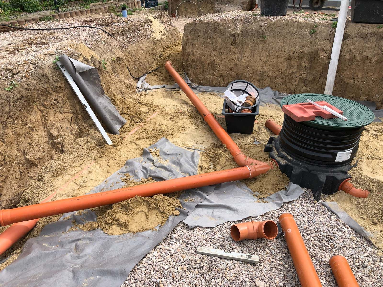 Orangefarbene Rohre und eine Versorgungsgrube auf einer schlammigen Baustelle mit Kies und abgedeckten Grabenabschnitten