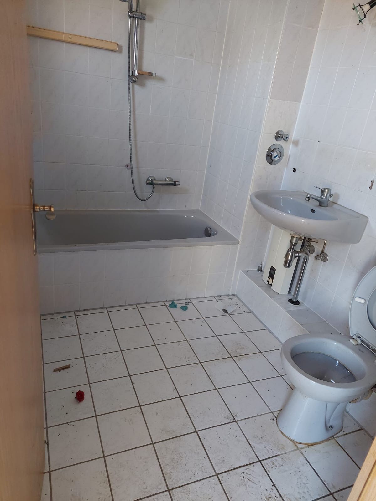 Kleines weißes Badezimmer mit Badewanne, wandmontiertem Waschbecken und Toilette auf gefliestem Boden.