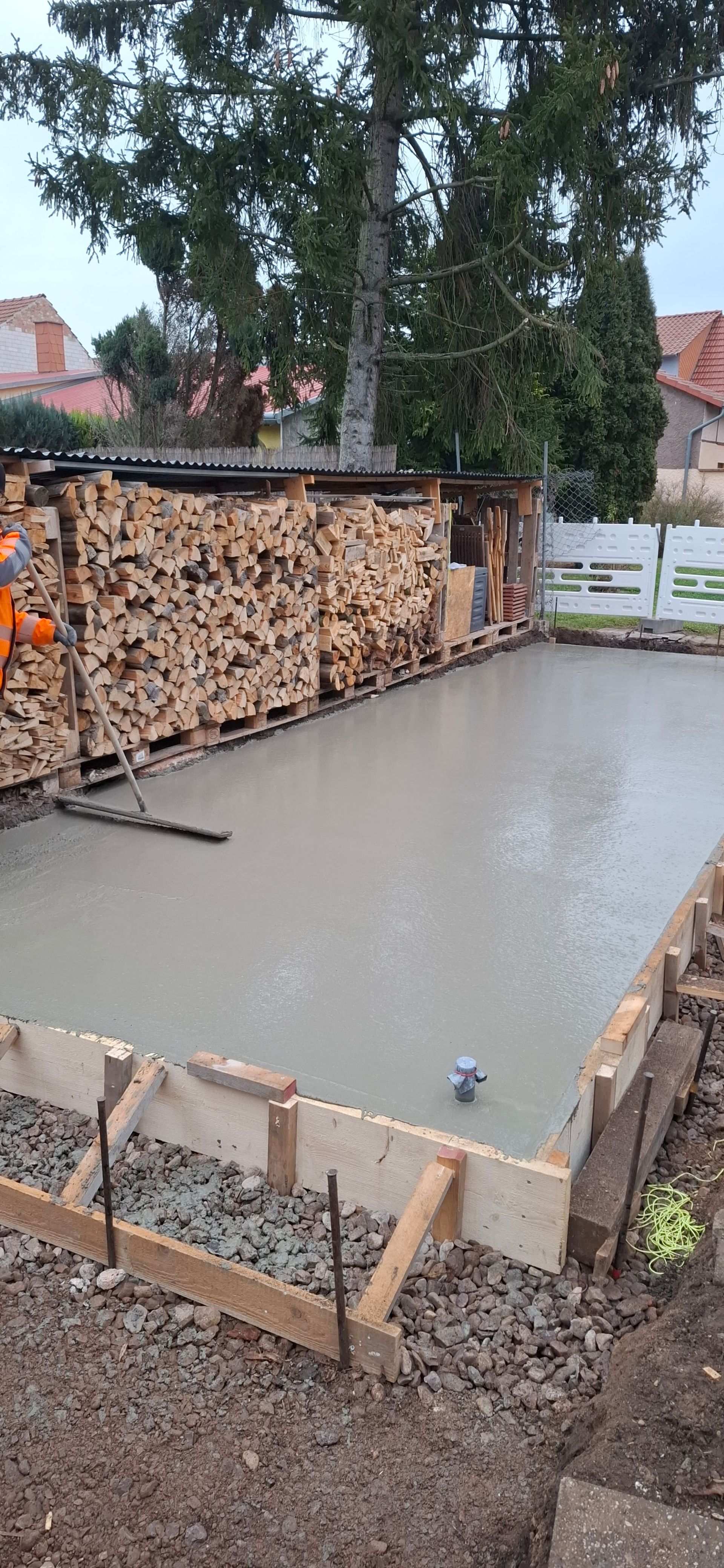 Frische Betonfundamentplatte mit Holzschalung und gestapeltem Holz im Hof