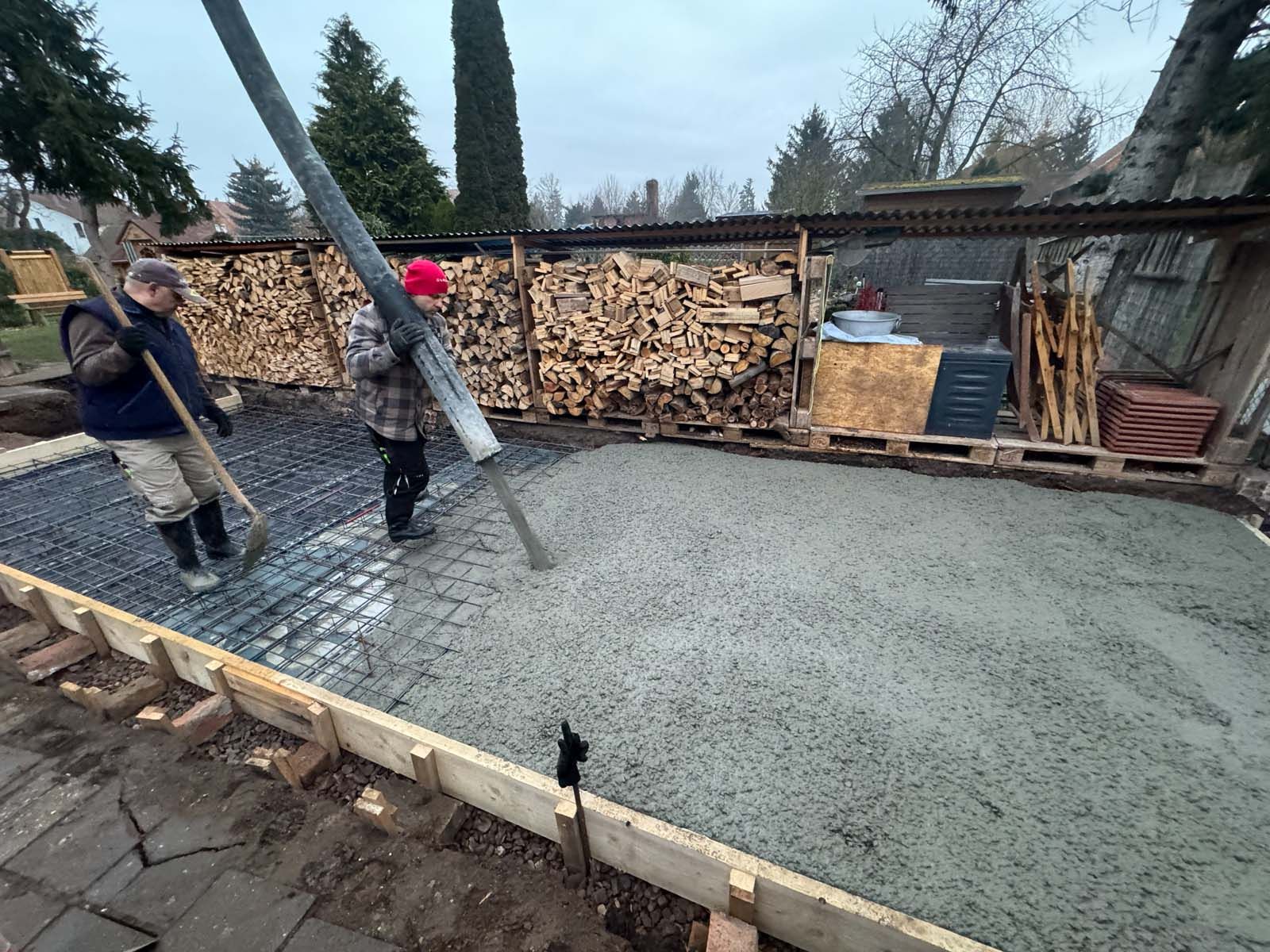 Arbeiter gießen eine Betonplatte neben gestapeltem Brennholz in einem matschigen Außenhof.