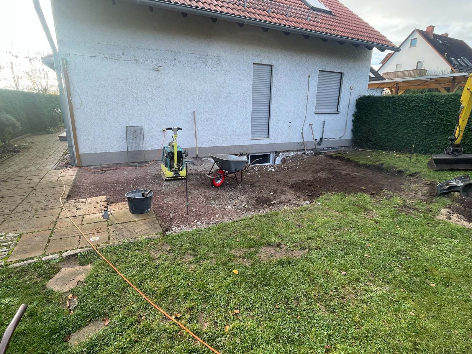 Hinterhof mit einem kleinen weißen Haus, gepflasterter Terrasse und einem unbefestigten Gartenbereich mit Werkzeugen und einer Schubkarre