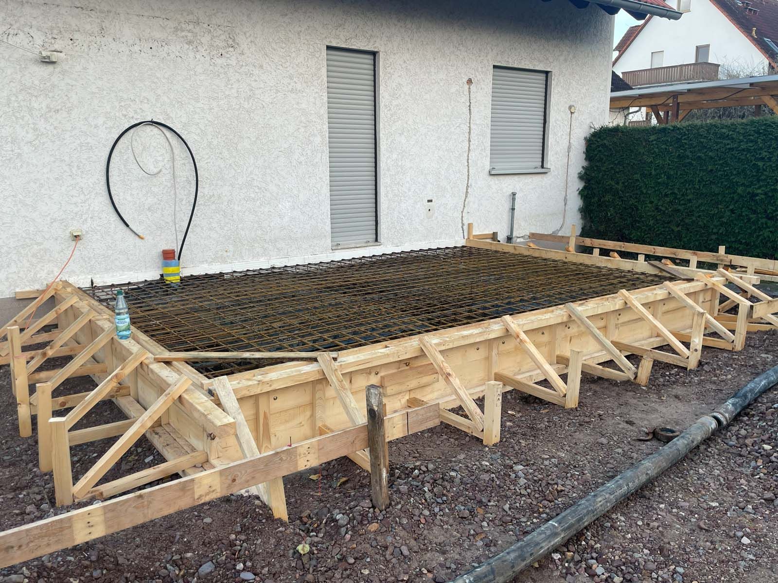 Schalung für eine Betonplatte im Hinterhof neben einem Haus, mit bereits vorhandener Holzrahmenkonstruktion und Bewehrungsgitter.