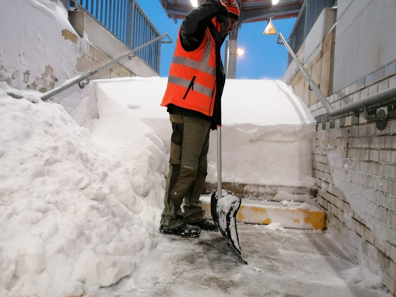 Ein Arbeiter in orangefarbener Weste schaufelt Schnee auf einer Außentreppe neben einem Gebäude.