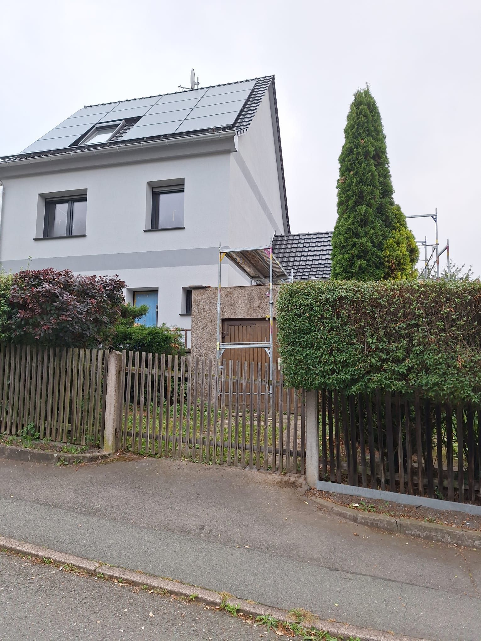 Graues zweistöckiges Haus mit schrägem Dach, umzäuntem Vorgarten und Hecke an einer ruhigen Straße.