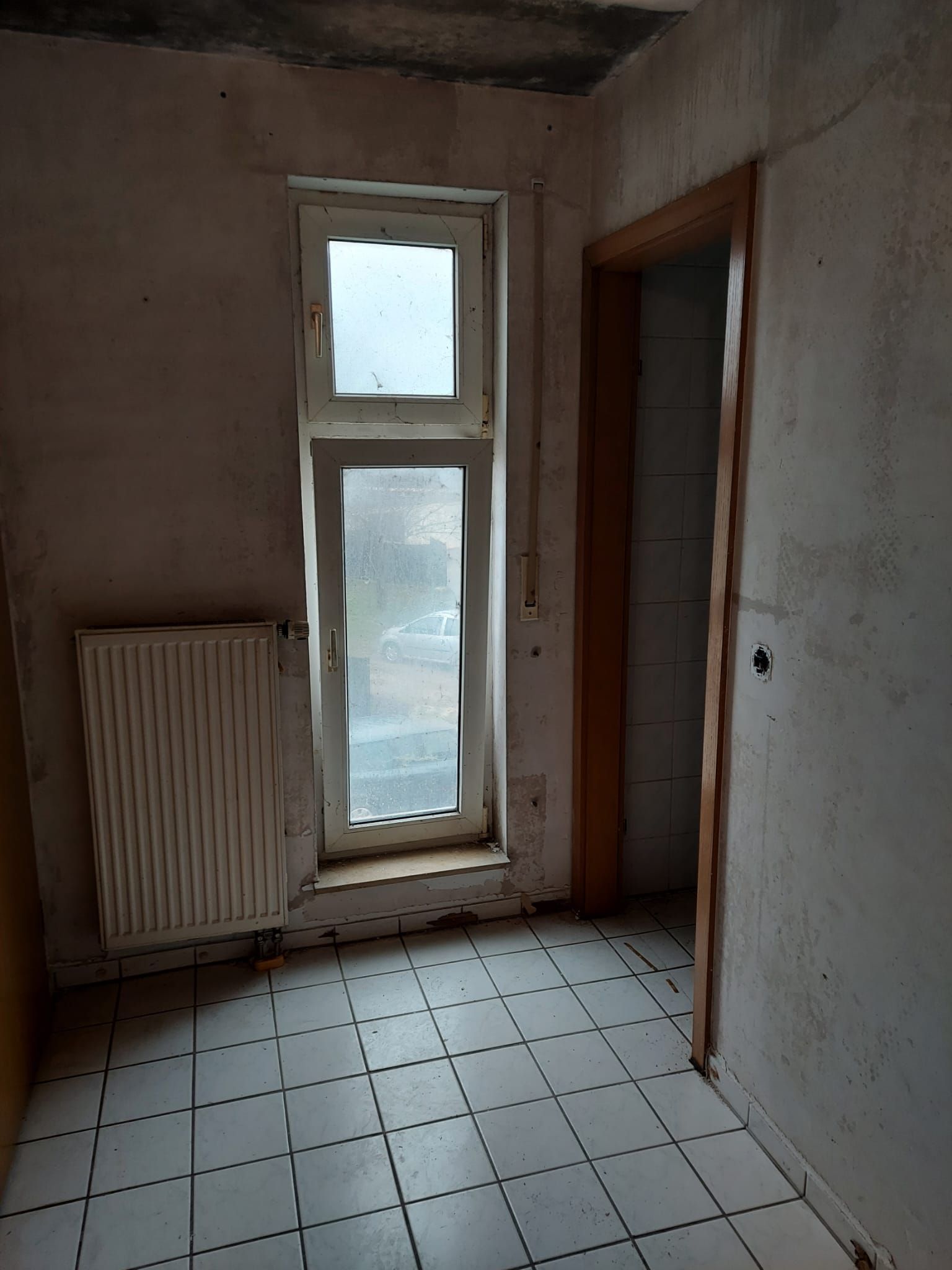 Leerer Flur mit weiß gefliestem Boden, Heizkörper und Milchglastür in einem abgenutzten Interieur.