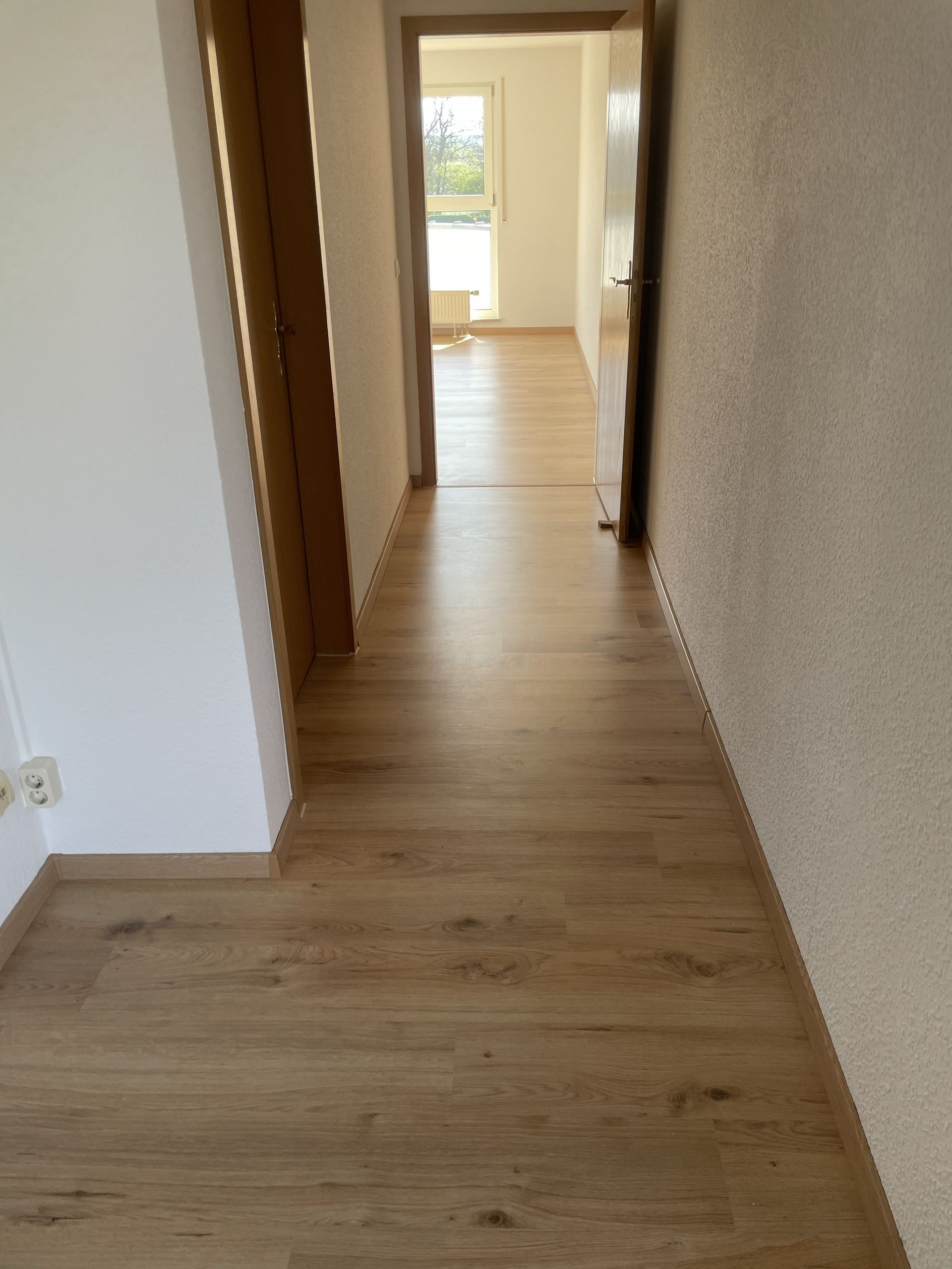 Ein schmaler Flur mit hellem Holzboden und weißen Wänden führt zu einer hellen Tür am Ende.