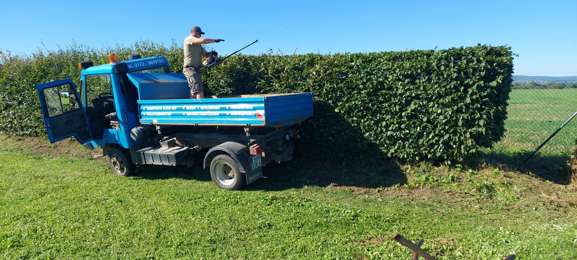 Ein blauer Geländewagen besprüht eine hohe grüne Hecke auf einem sonnigen Feld.