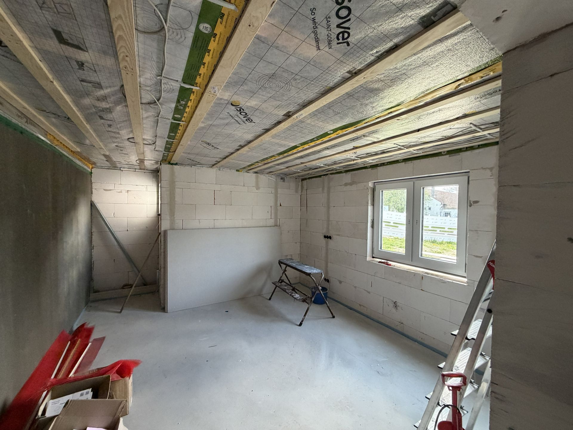 Unfertiger Raum mit freiliegender Dämmung, Gipskartonwänden, Fensterlicht und Bauwerkzeugen auf dem Boden.