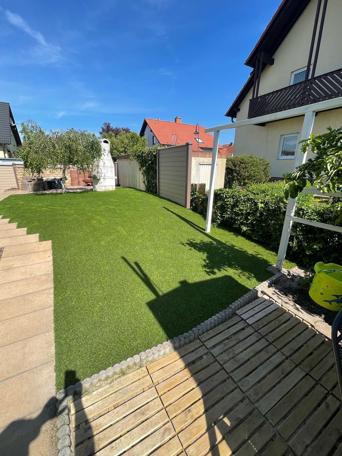 Sonniger Hinterhof mit grünem Rasen, gepflasterter Terrasse und weißen Hauswänden unter blauem Himmel
