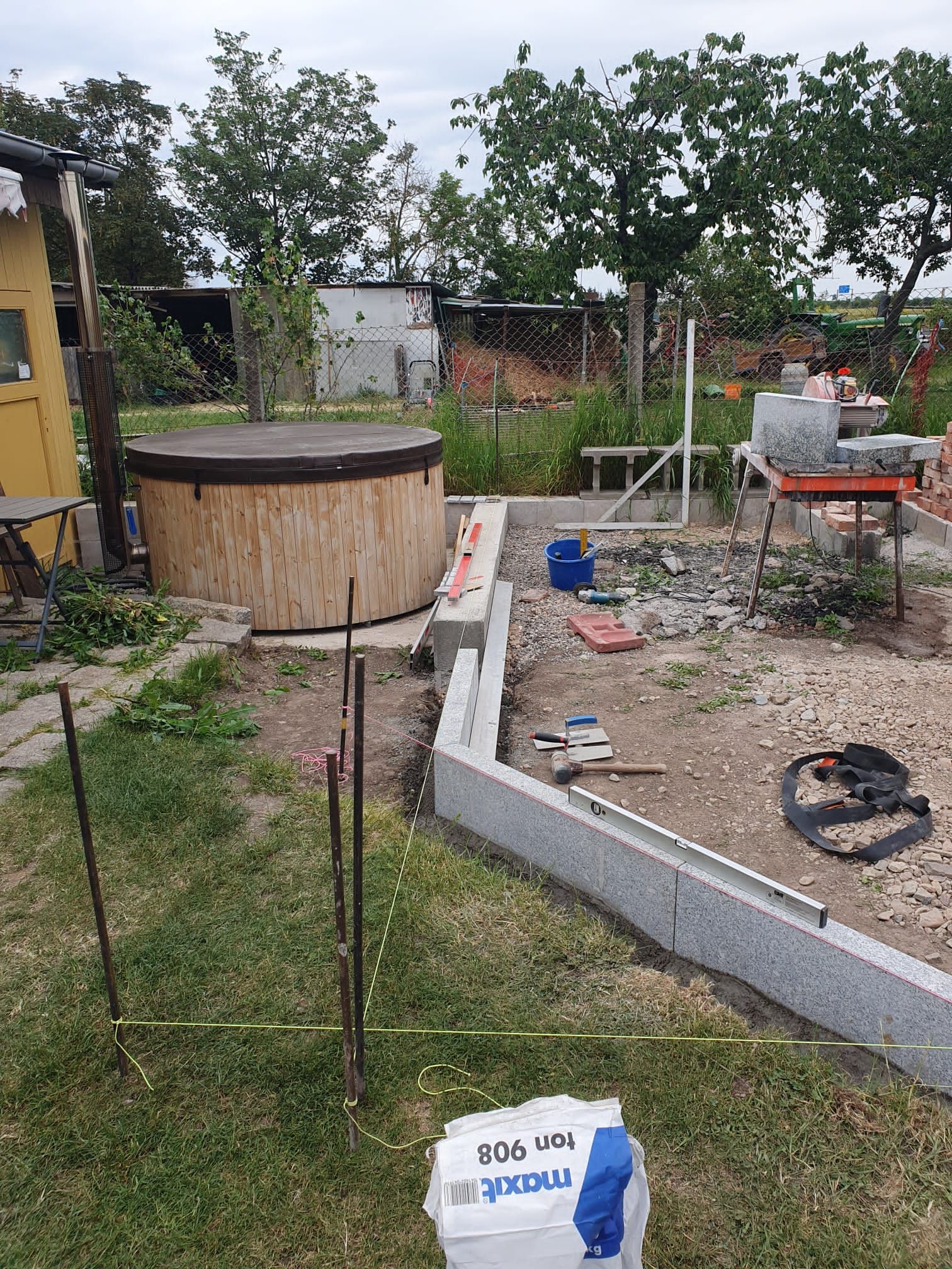 Hinterhof-Baustelle mit erhöhtem Betonrand, Holzfass, Werkzeugen und verstreuten Materialien.