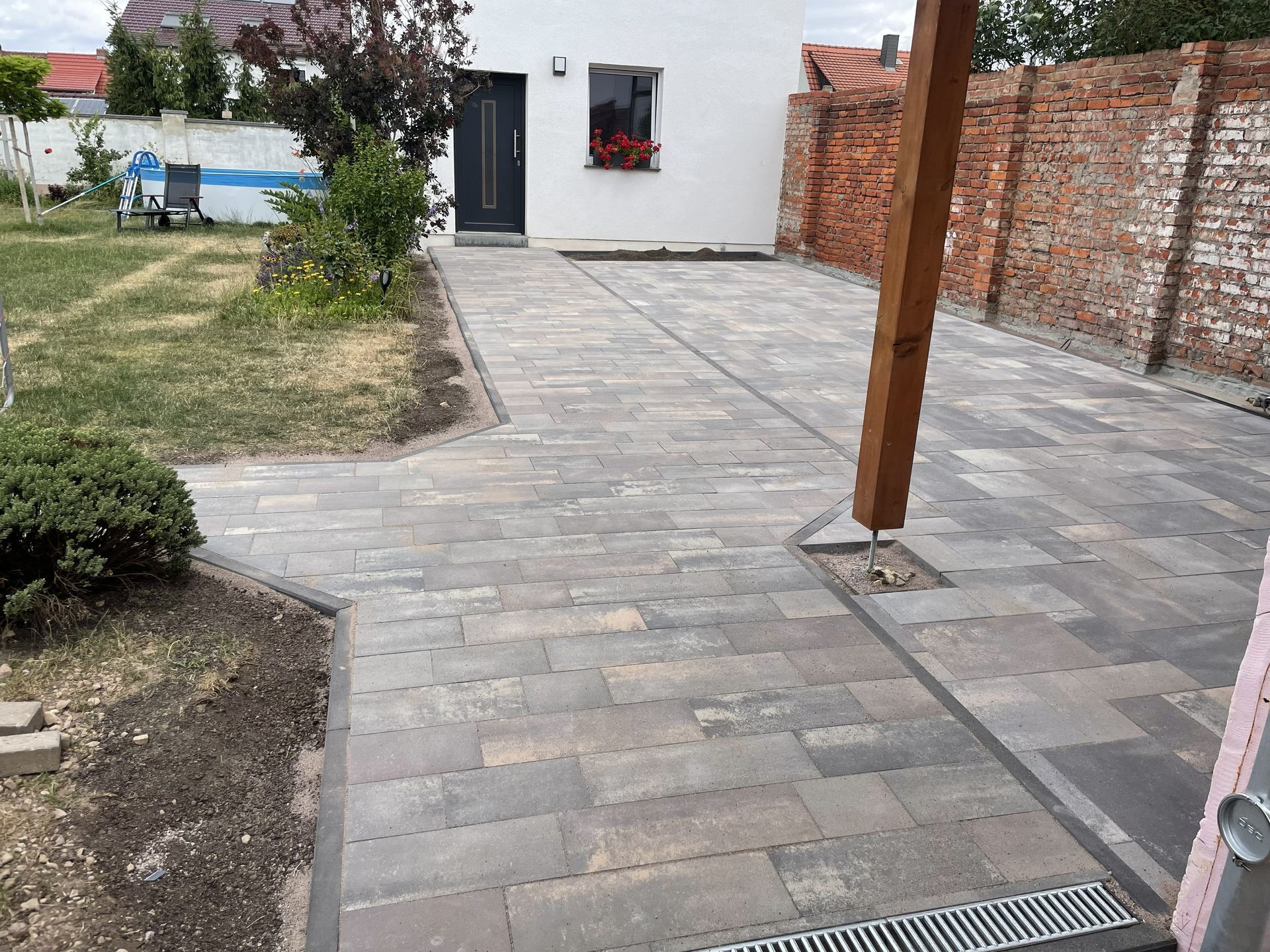 Gepflasterte Terrasse im Hinterhof mit Holzpfosten, Backsteinmauer, kleinem Rasen und einer weißen Haustür im Hintergrund