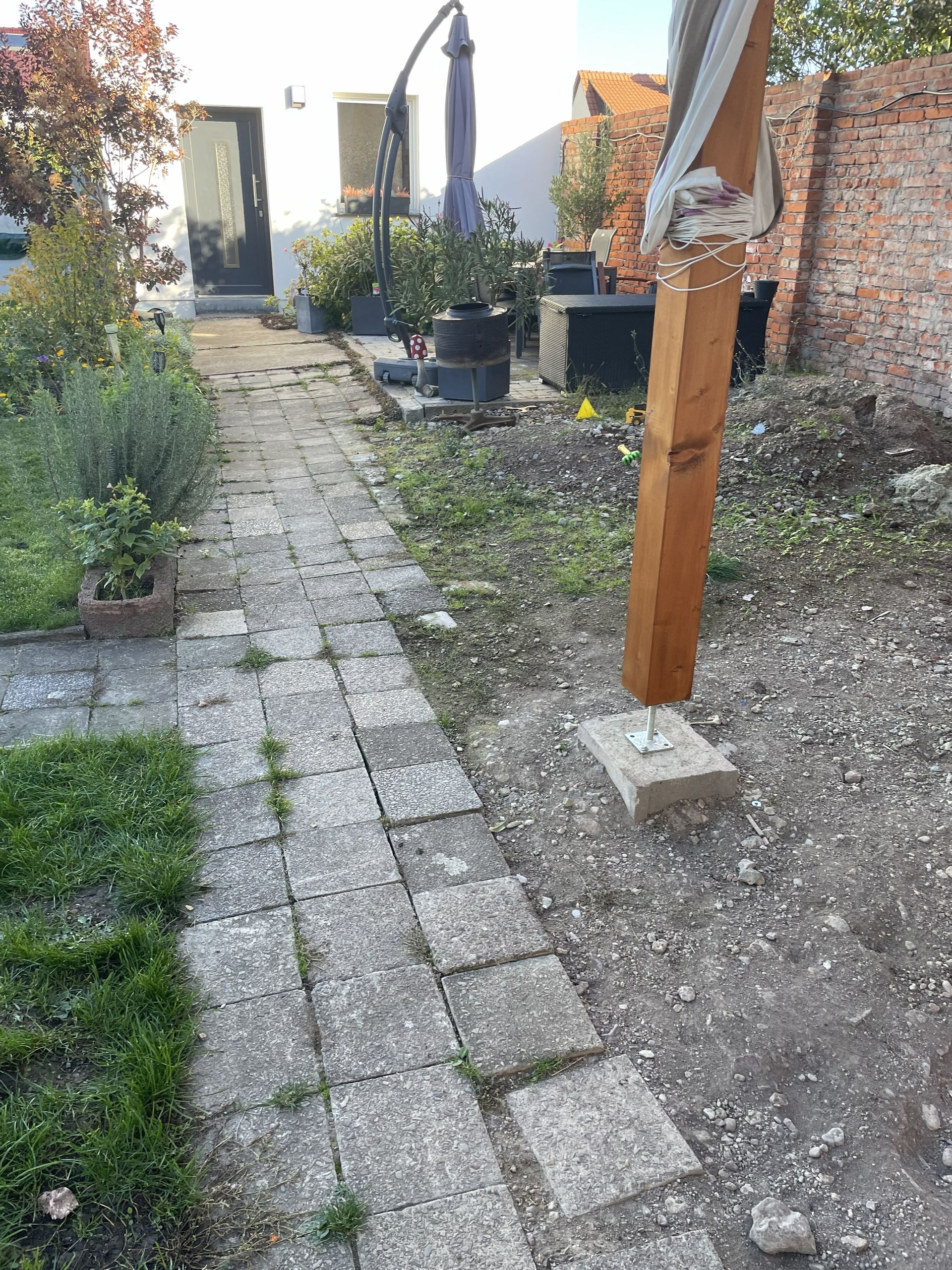 Ein schmaler Steinpfad neben einer Backsteinmauer führt zu einem Gartentor mit Topfpflanzen und einem Holzpfosten.