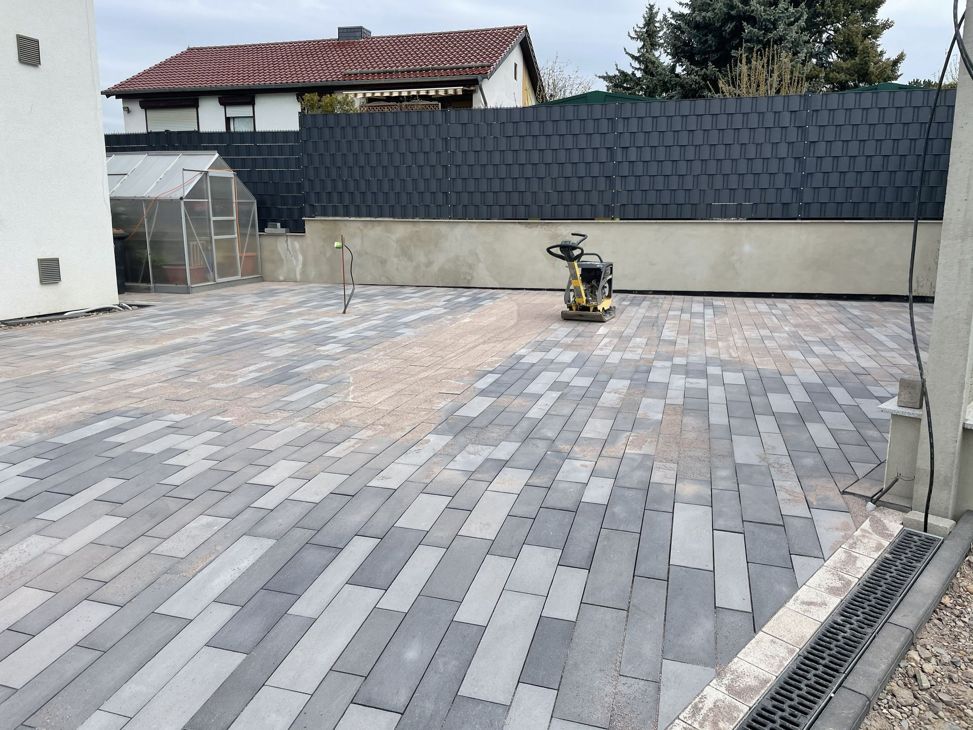 Eine gepflasterte Terrasse befindet sich im Bau; in der Mitte steht eine Rüttelplatte, im Hintergrund ein dunkler Zaun.