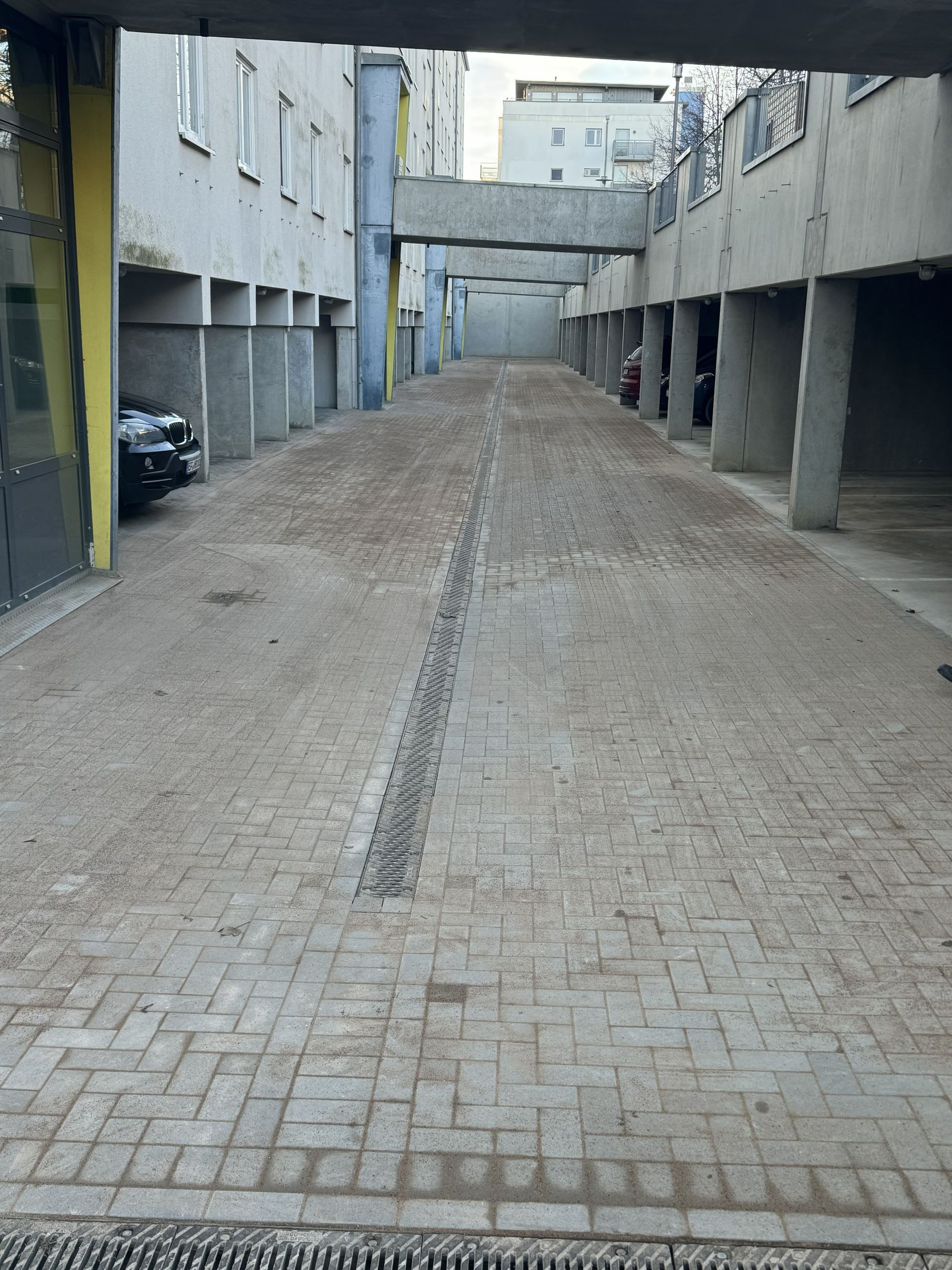 Leere, gepflasterte Gasse zwischen grauen Gebäuden mit geparkten Autos und überdachten Fußgängerbrücken