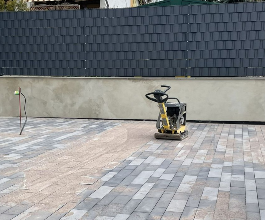 Eine gepflasterte Terrasse befindet sich im Bau; eine Rüttelplatte steht auf grauen und beigen Steinplatten in der Nähe einer Mauer.