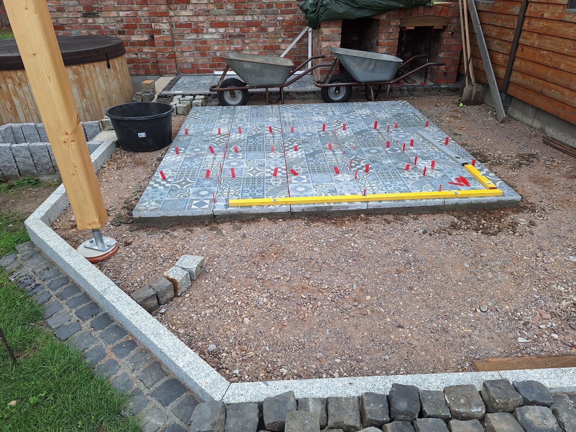 Hinterhof-Terrassenbereich mit Kies, Randsteinen, Schubkarre und einer überdachten rechteckigen Platte neben einer Ziegelmauer.