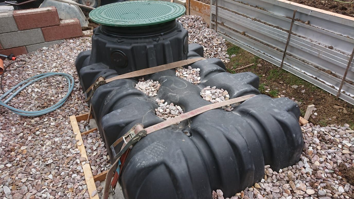Schwarzer unterirdischer Wassertank mit grünem Deckel, festgeschnallt in Kies neben einem Zaun und einem Schlauch.