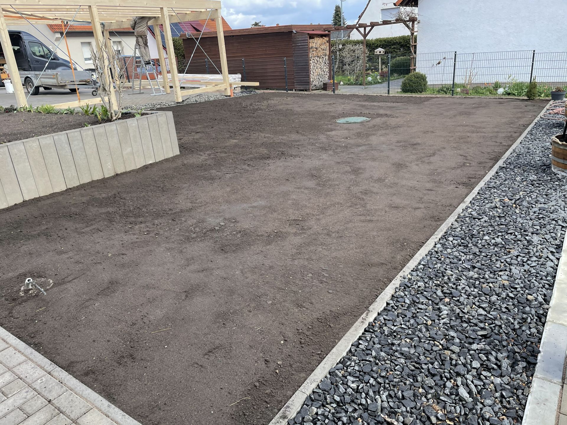 Ein unbebautes Grundstück, das von Kies und Pflastersteinen begrenzt wird, im Hintergrund ein kleiner Schuppen und ein Zaun.