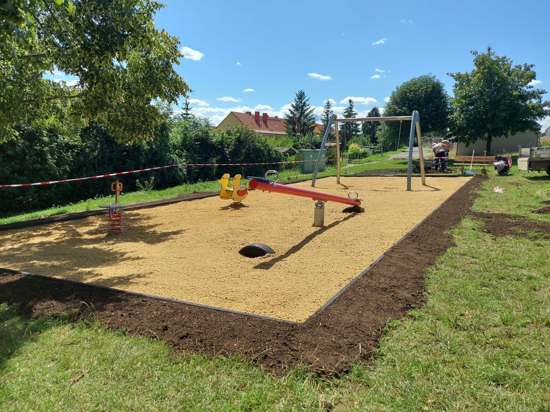 Außenspielplatz mit Sandspielfläche, Balancierbalken und einem Klettergerüst aus Metall, umgeben von Gras und Bäumen.