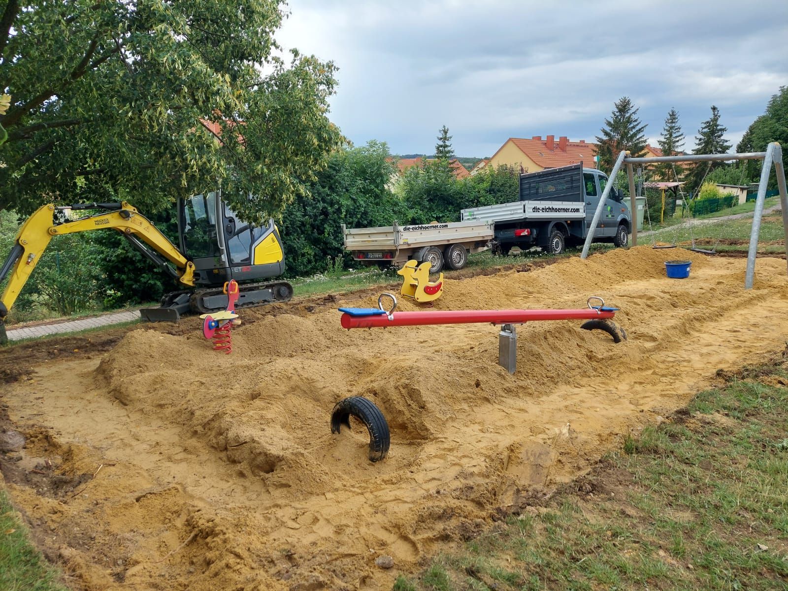 Spielbereich auf einer Baustelle mit gelbem Bagger, rotem Balancierbalken und Lastwagen auf einem Sandhügel im Freien.