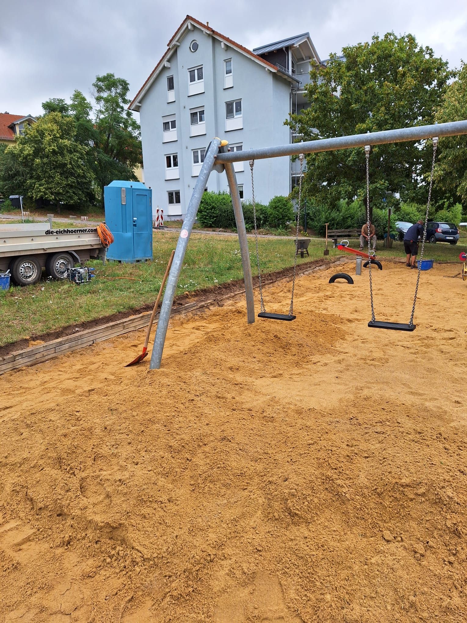 Blaue Schaukel auf Holzschnitzeln auf einem Spielplatz neben einem hellblauen Haus und Bäumen nach dem Regen
