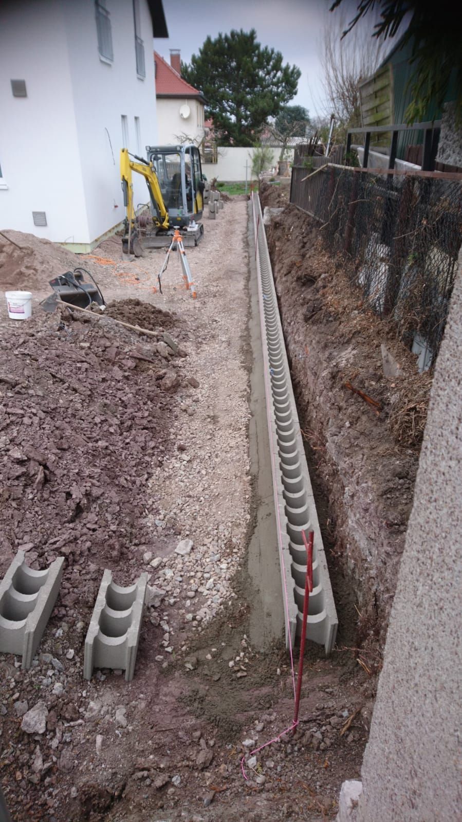 Schmaler Graben neben einem Haus mit Betonkabelkanal und Bauschutt entlang eines Zauns.