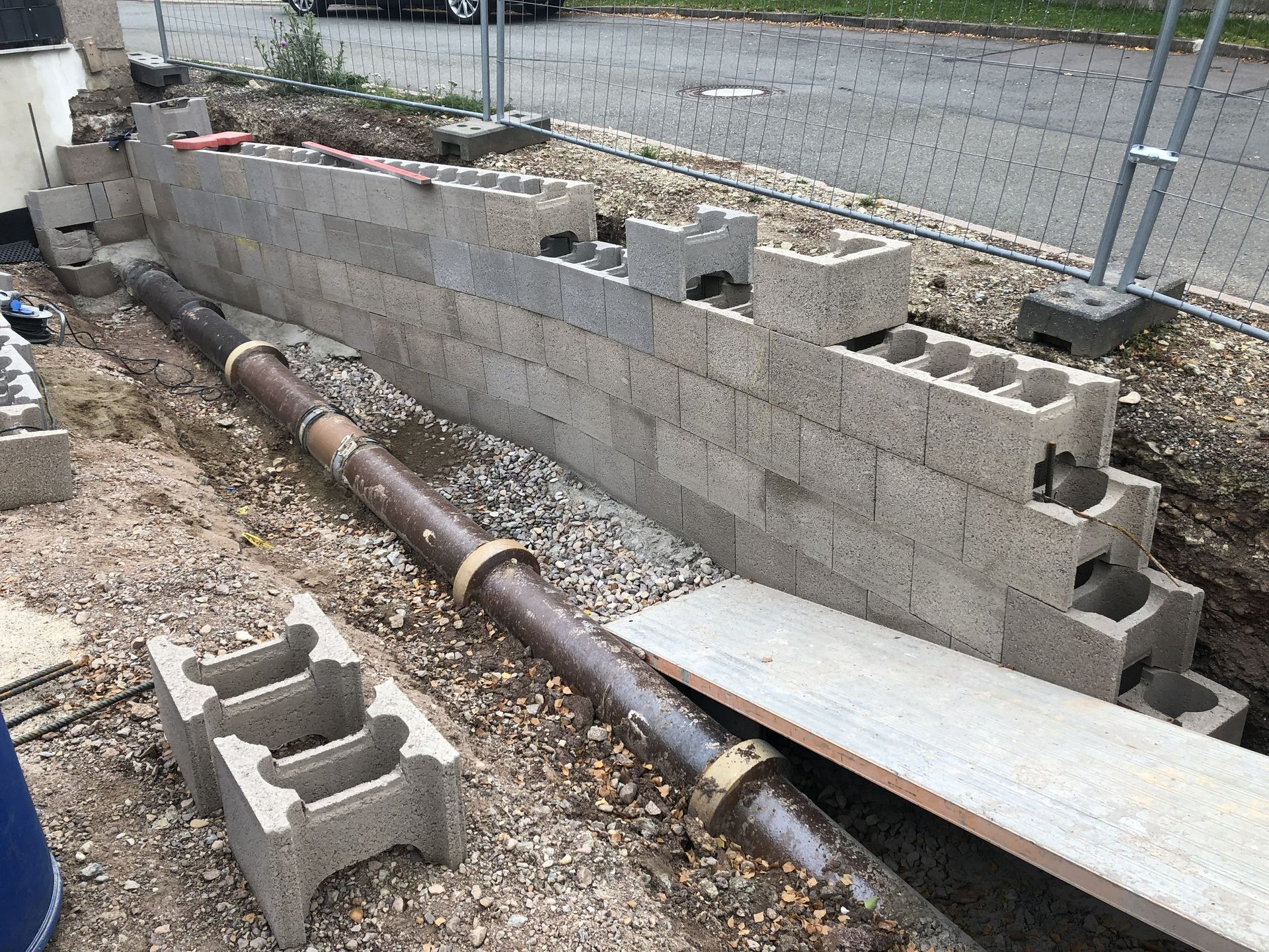 Baustelle mit gestapelten Betonblöcken und einem verrosteten Rohr neben einem Kiesgraben und einem Metallzaun