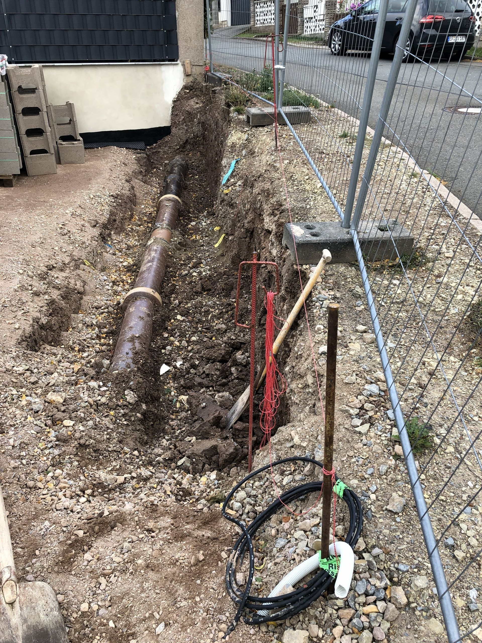 Ausgehobene Erde neben einem Maschendrahtzaun, davor freiliegende Rohre, Kabel und Bauschutt vor einem Gebäude.