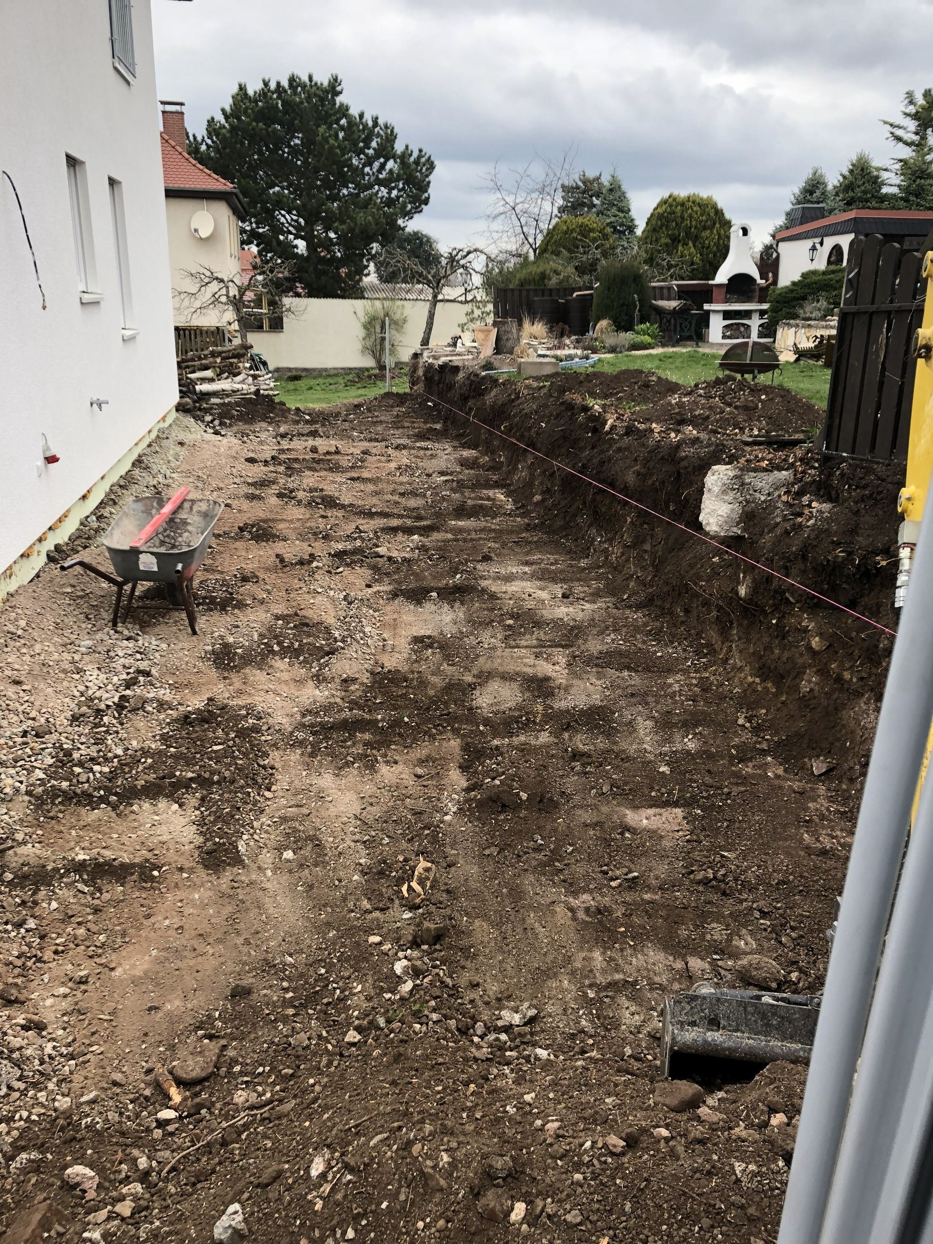 Ein matschiger Hinterhofweg neben einem weißen Haus, mit verstreutem Schmutz, kleinen Pflanzen und bewölktem Himmel.