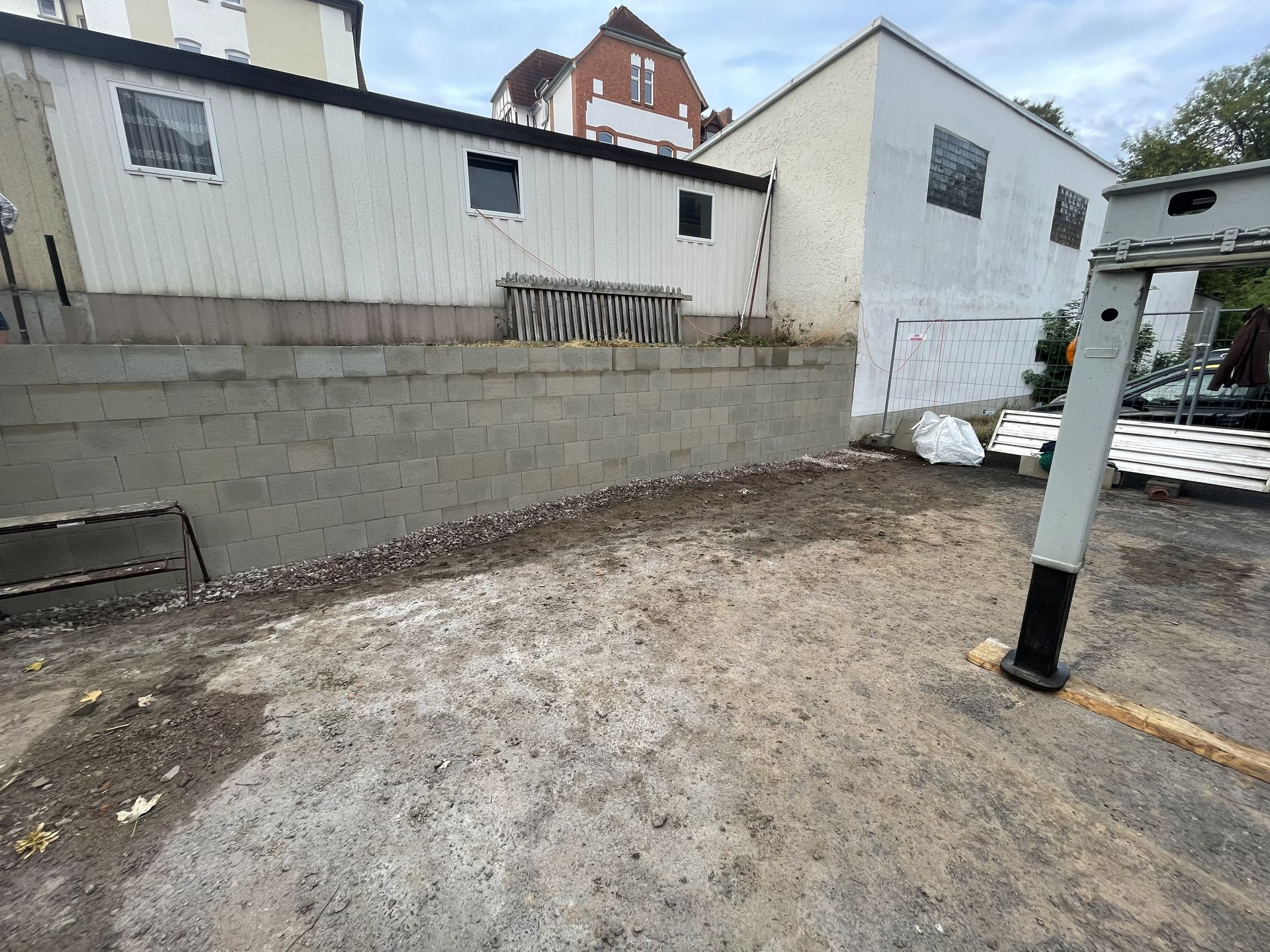 Kleiner unbefestigter Platz neben einem weißen Gebäude und einer Betonmauer, mit verstreuten Materialien und einem schwarzen Pfosten.