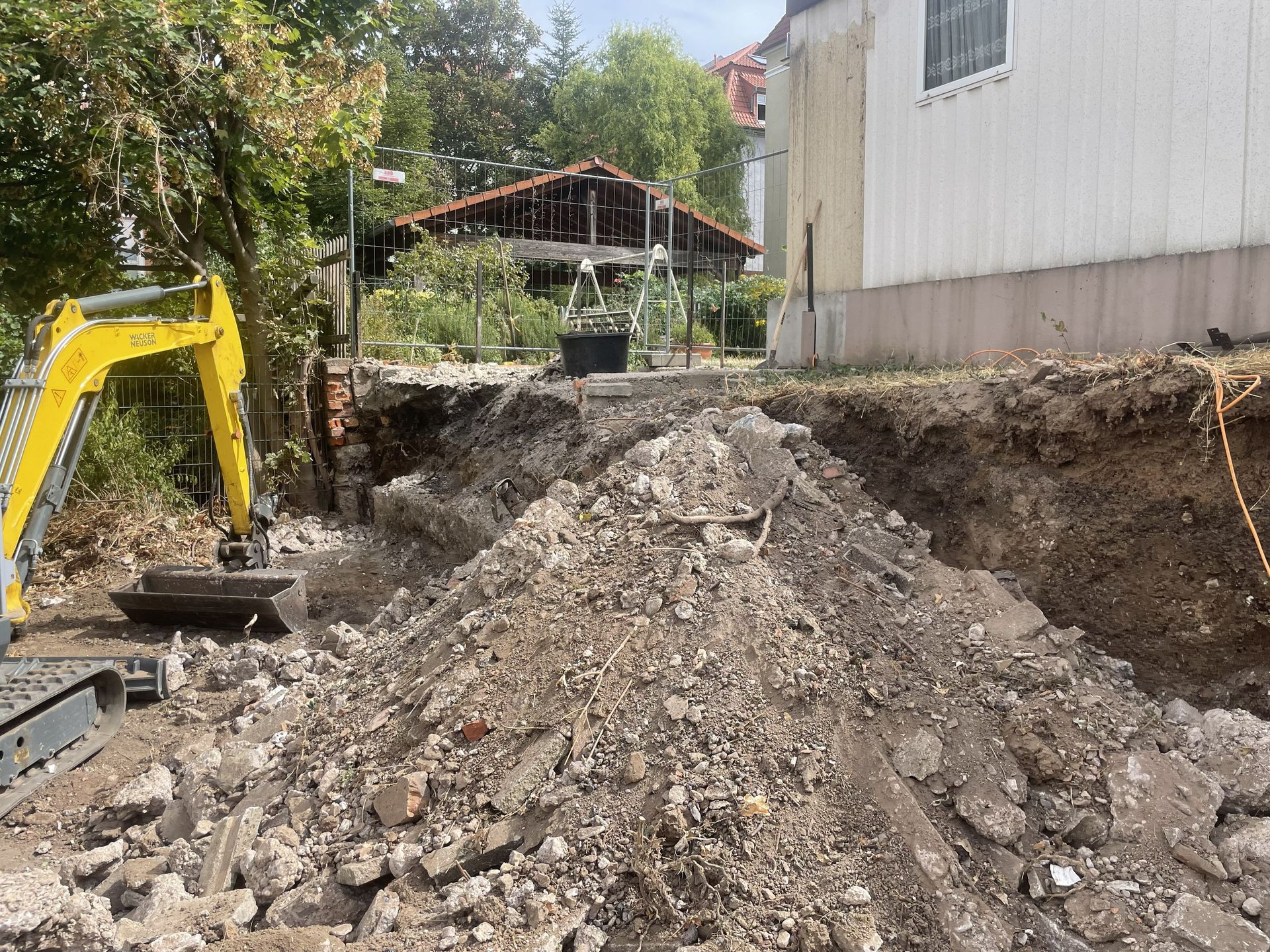 Ein Bagger hebt neben einem Haus einen großen Graben aus; im Vordergrund ist ein Haufen Erde und Bauschutt zu sehen.