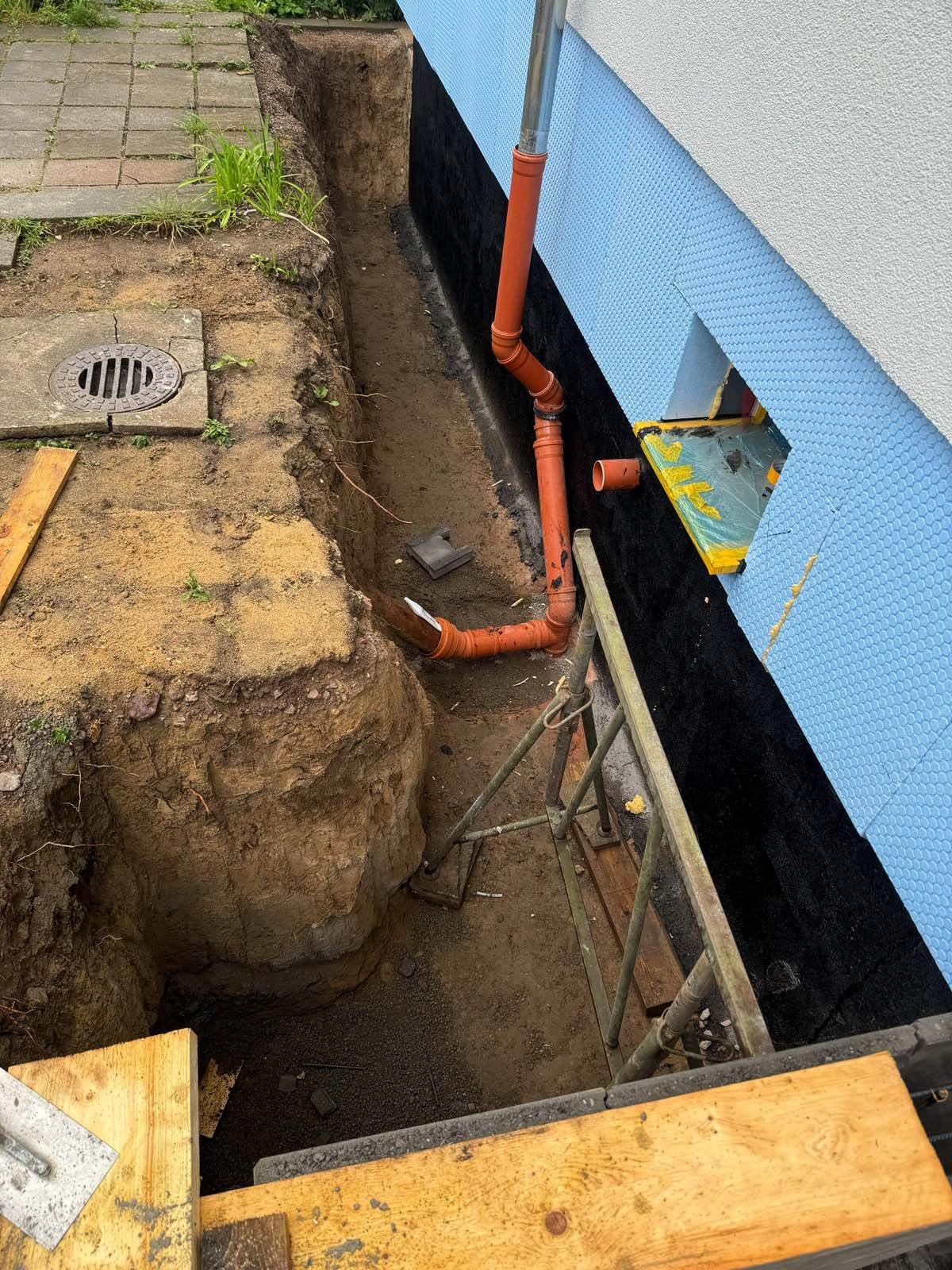 Freiluft-Aushubarbeiten neben einem blauen Gebäude mit freiliegenden orangefarbenen Rohren und einem Graben im Boden