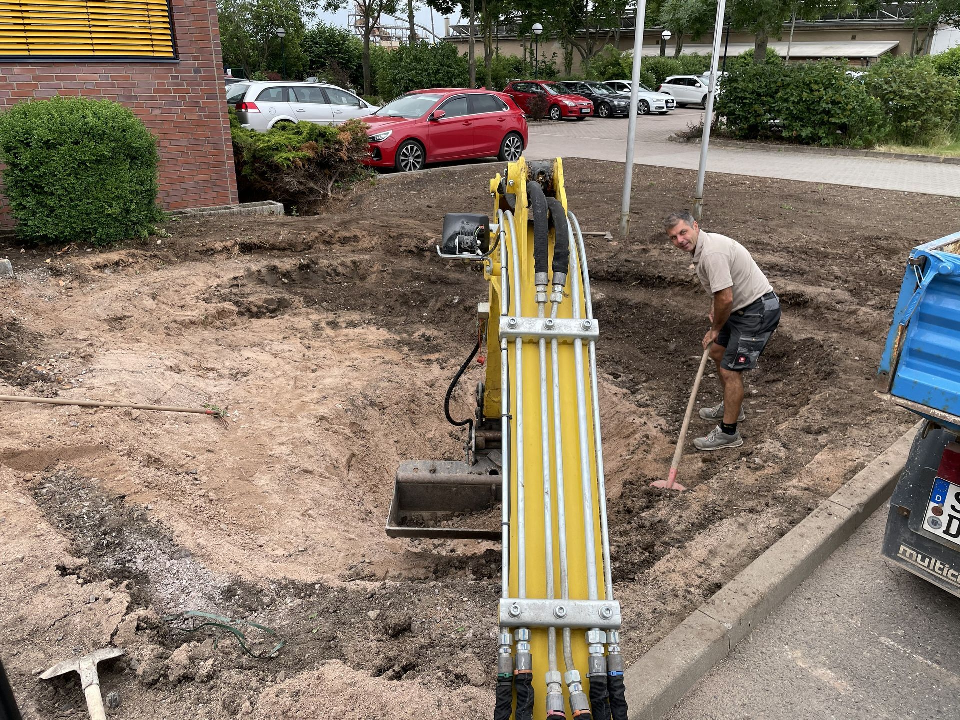 Ein Bagger gräbt einen Graben neben einem Bordstein, während ein Arbeiter Erde in der Nähe geparkter Autos wegräumt.