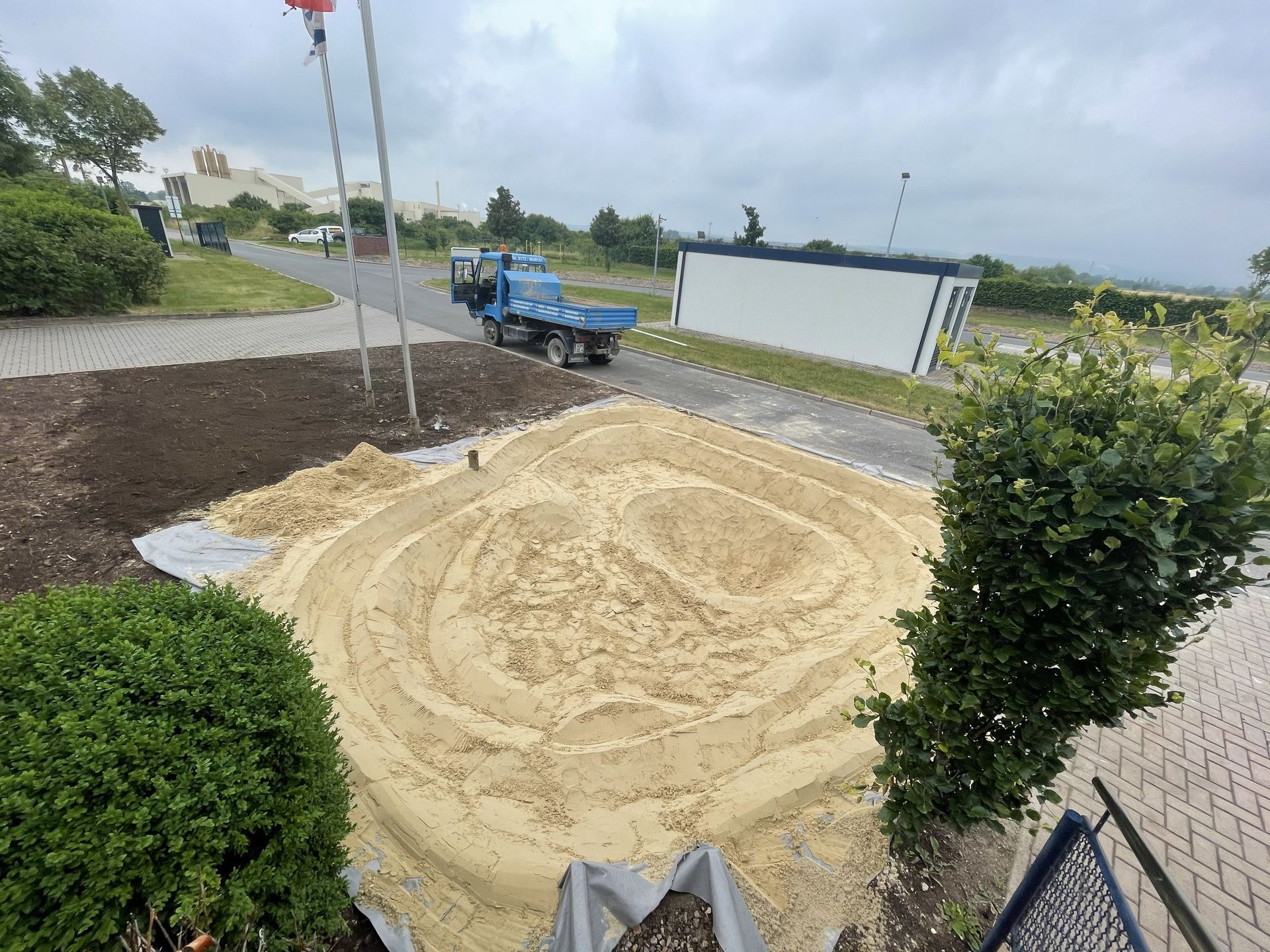 Sand verteilt sich in einem Hof ​​neben einem blauen Lastwagen und einer weißen Mauer unter bewölktem Himmel.