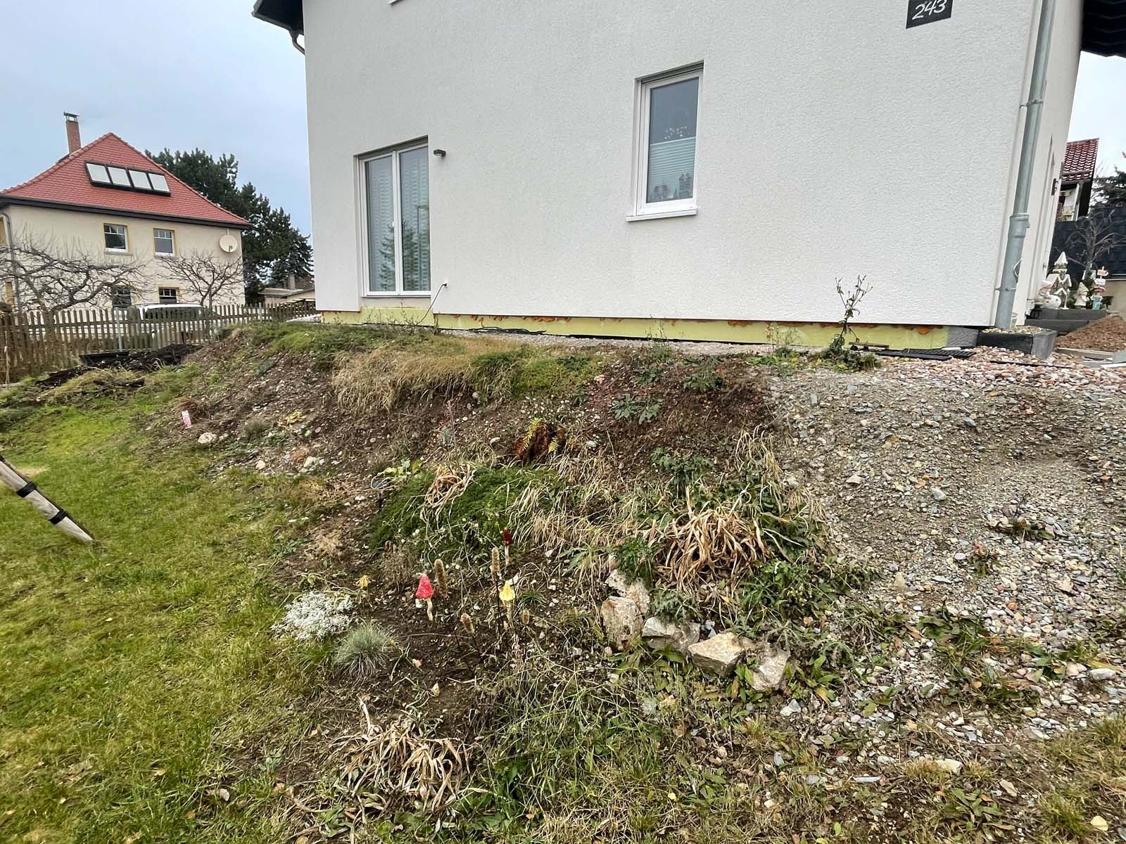 Hinterhof mit grasbewachsenem Hang und Kies neben einem weißen Haus, im Hintergrund die Häuser der Nachbarn mit roten Dächern.