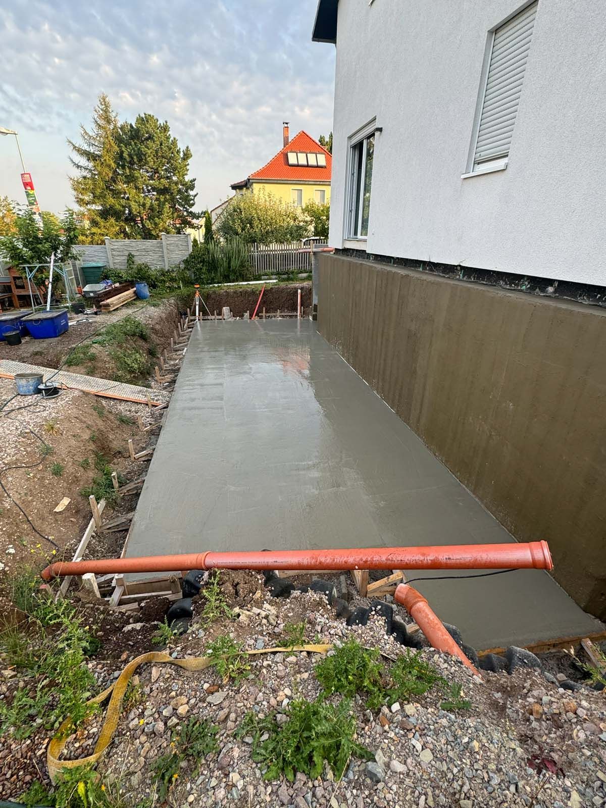 Eine frische Betonplatte neben einem Haus, im Vordergrund ein rotes Rohr und drumherum ein Kiesplatz.