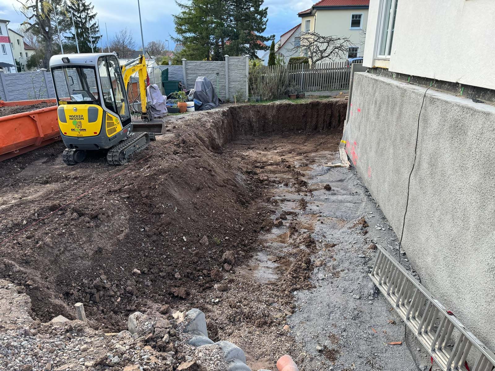 Ein Bagger hebt einen Graben neben einem Haus aus, entlang des Fundaments ist Erde aufgeschüttet.