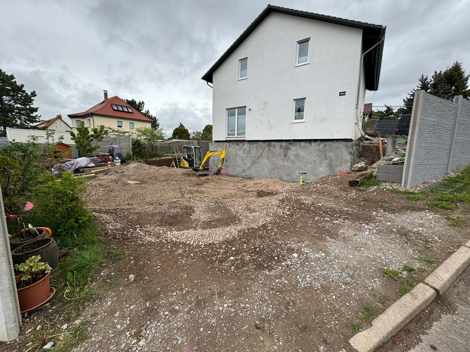 Ein unbewachsener Hof neben einem weißen Haus, mit Bordstein, Blumen und einem Haus mit rotem Dach im Hintergrund