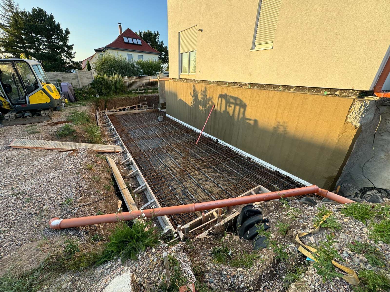 Baustelle neben einem gelben Haus mit Stahlbetonfundament, roten Rohren und Kiesgraben